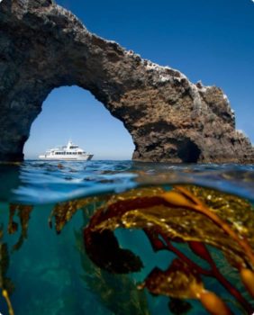 yacht under a rock arch, half image is under clear water with seaweed below