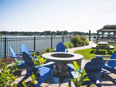 Stone fire pit surrounded by blue Adirondack chairs overlooking the water and a gazebo in the background